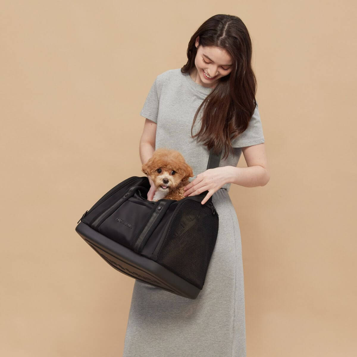 A dog peeking out of the top opening of the Pups and Bubs Onboard Travel Carrier with a woman demonstrating how it looks like when slung across the body.