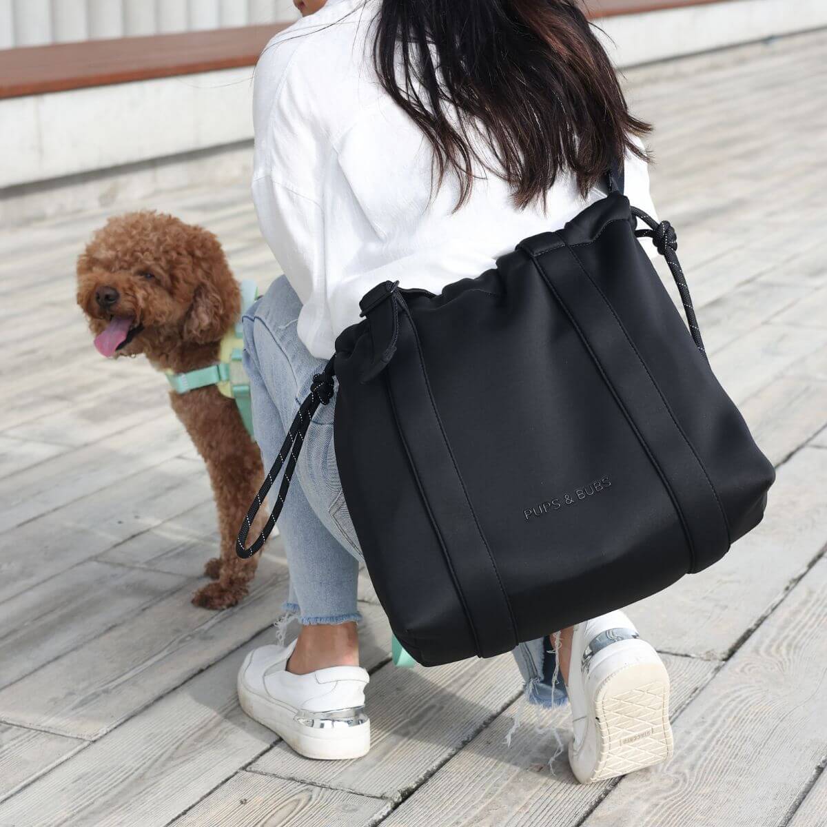 A woman wearing the Pups and Bubs All Day Chill Bag in Black as a crossbody bag, with a brown poodle in front of her.