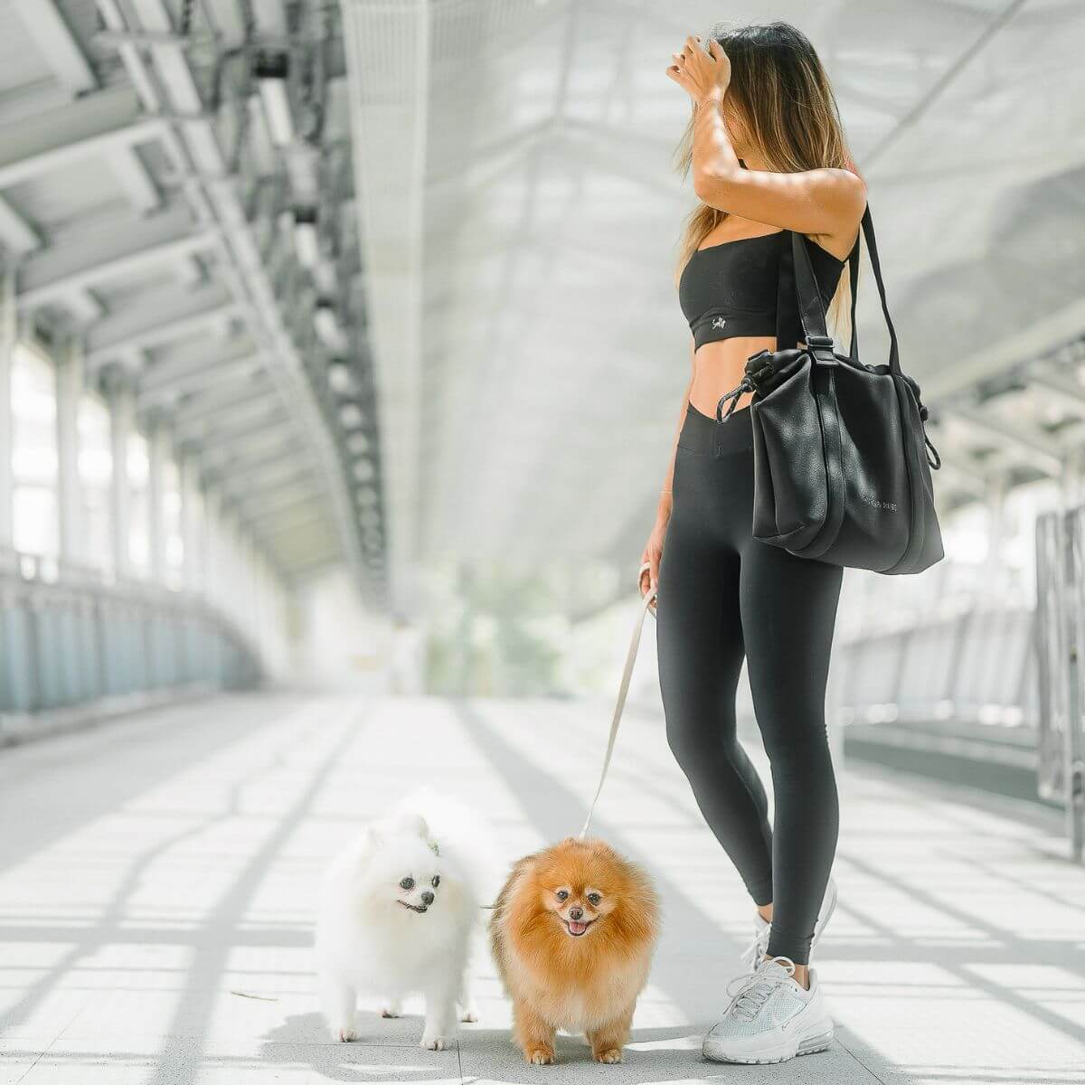 A woman in activewear carrying the Pups and Bubs All Day Chill Bag in Black on one shoulder while leashing two pomeranians, one white and one brown.