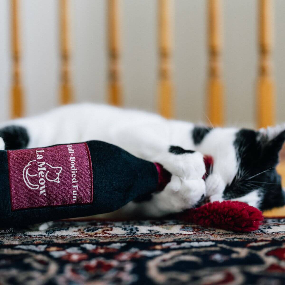 A black and white cat lying on a carpet on its side with its paws gripping onto the Play Pet Feline Frenzy Catbernet Sauv Kicker.