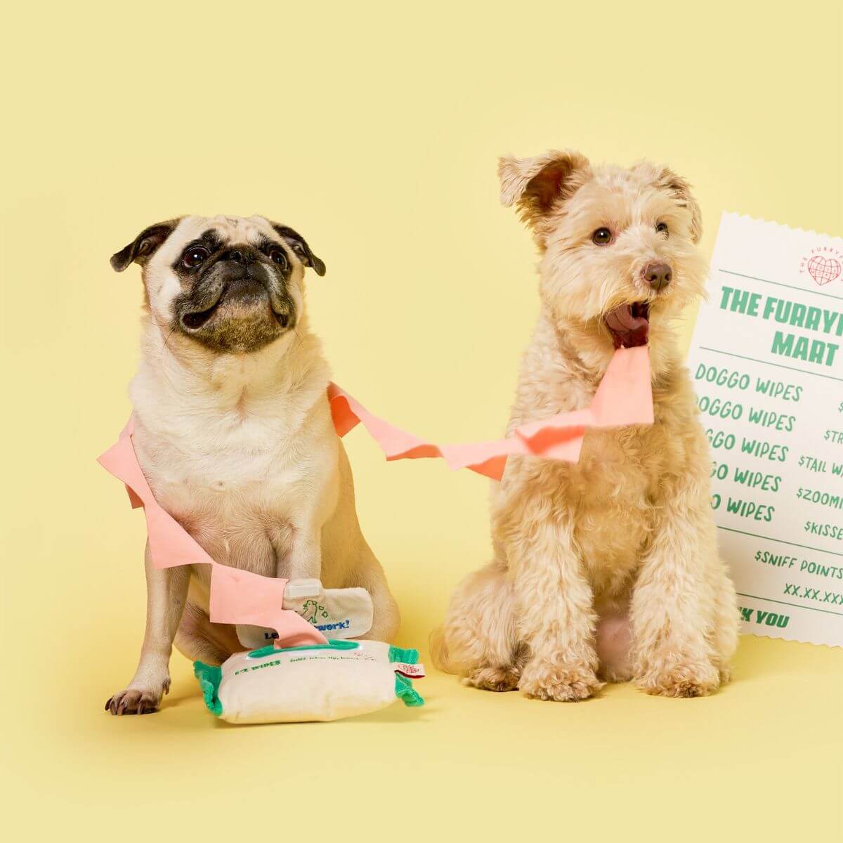 2 dogs sitting on the floor against a yellow background with the the furryfolks doggo wipe nosework toy draped around them.
