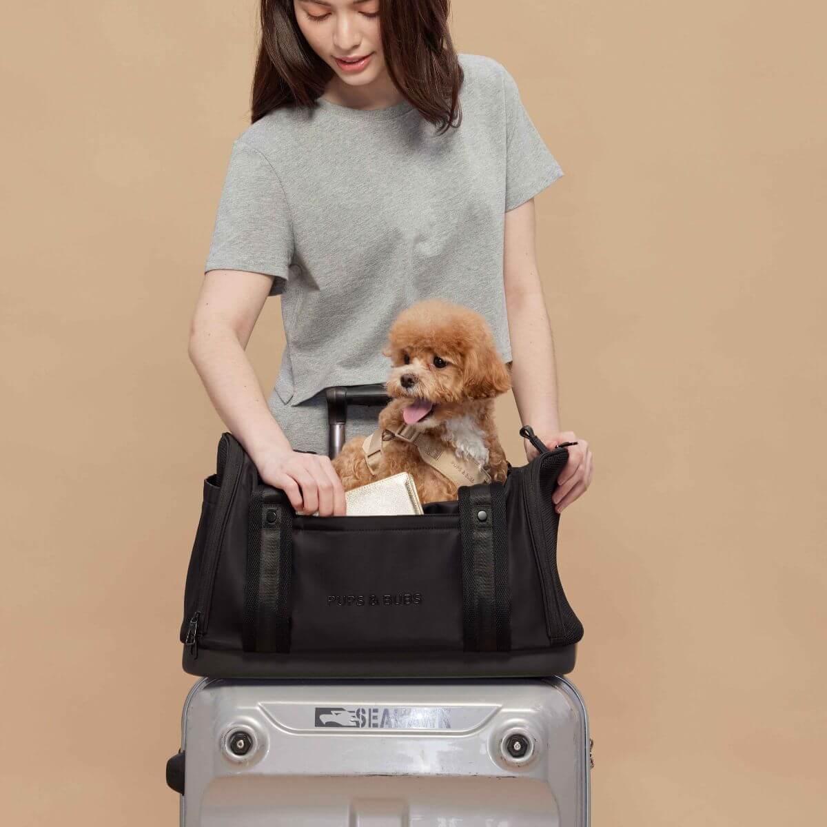 A dog peeking out of the top opening of the Pups and Bubs Onboard Travel Carrier with a woman demonstrating front pockets for storage.