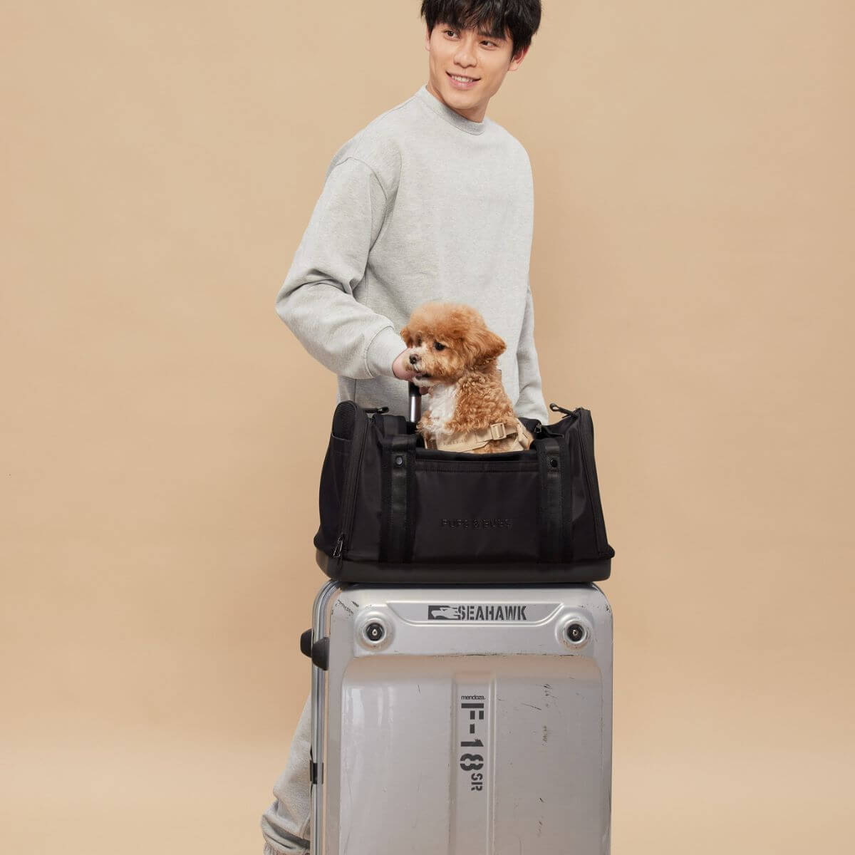 A dog peeking out of the top opening of the Pups and Bubs Onboard Travel Carrier with a man demonstrating luggage slot for easy transport.