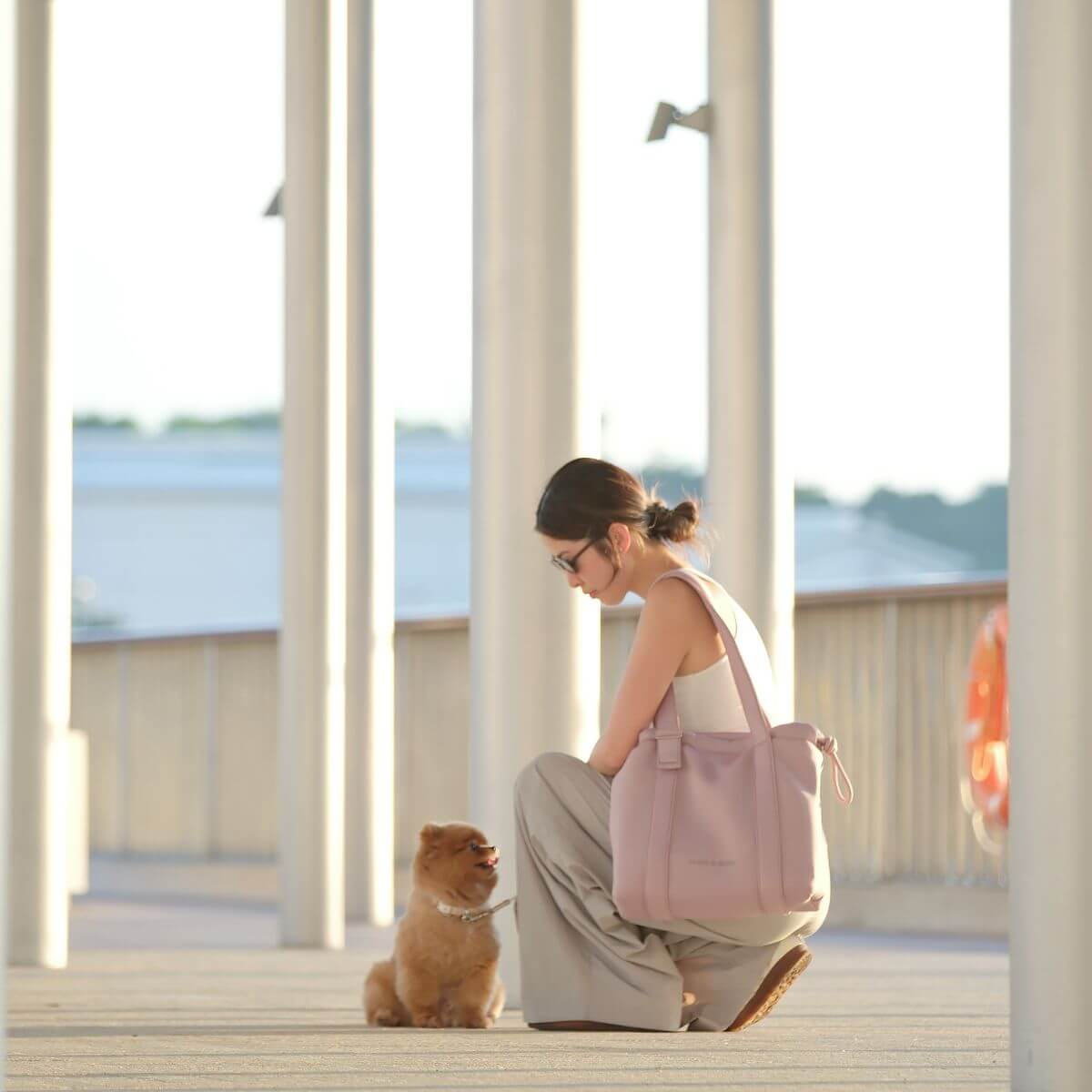 A woman squatting next to a brown pomeranian while carrying the Pups and Bubs All Day Chill Bag in Rose on one shoulder.