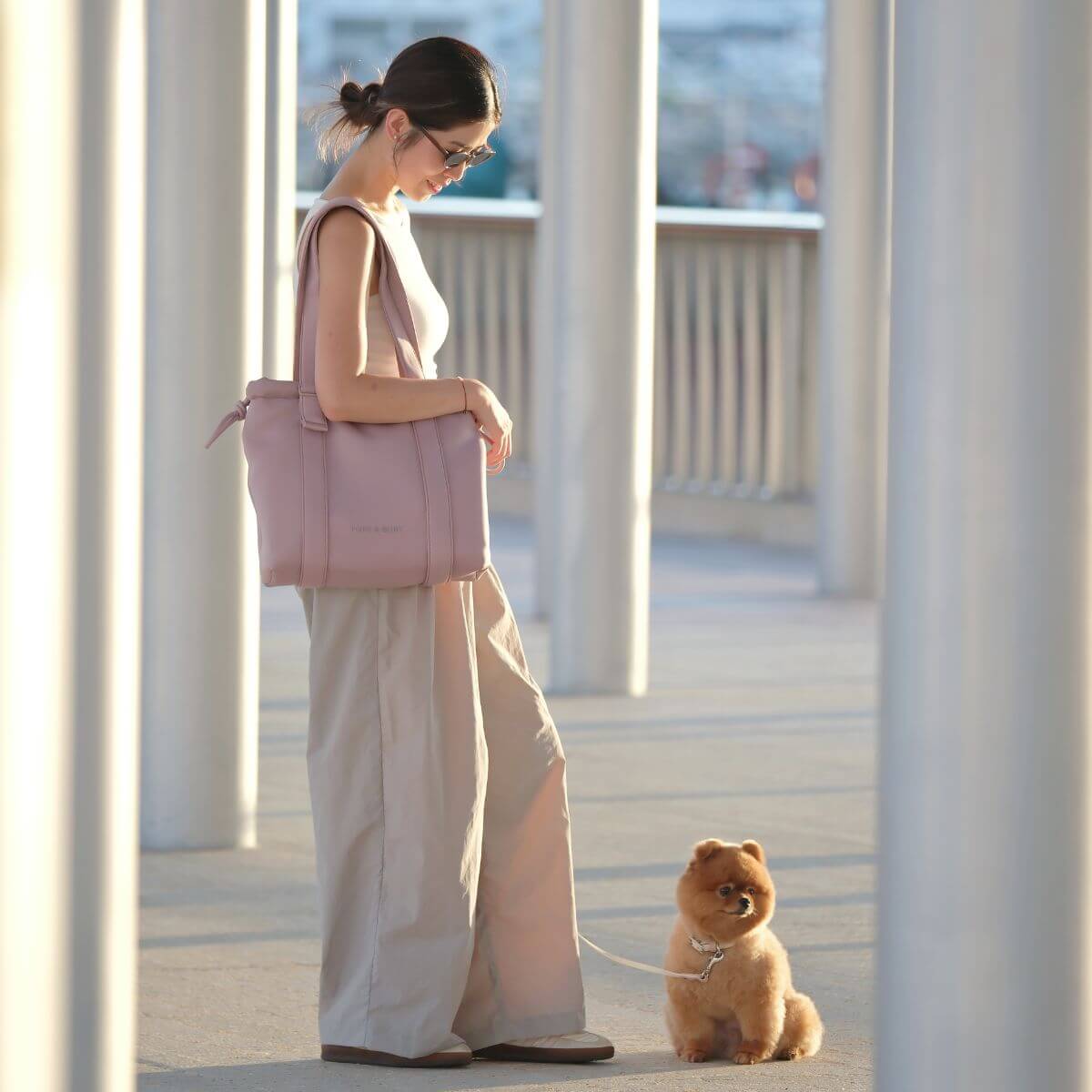 A woman carrying the Pups and Bubs All Day Chill Bag in Rose and a brown pomeranian on the ground pose for the photo.
