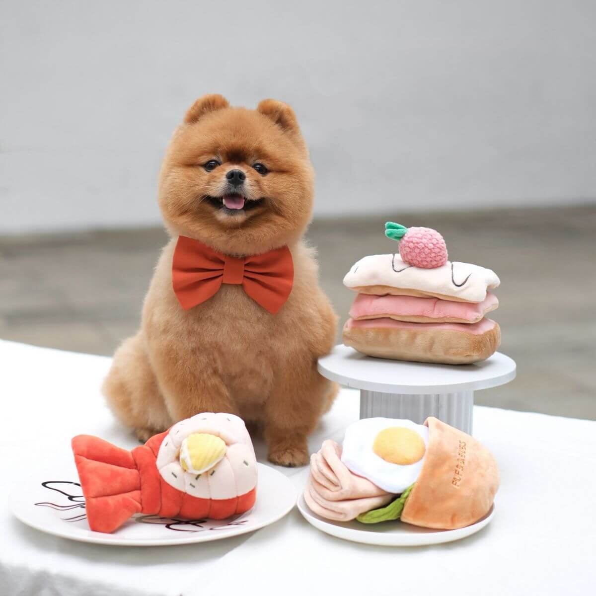 A pomeranian with a red bow tie posing with the Pups and Bubs Pita Pocket, Lobster, and Strawberry Millie-Feullie Nosework Toys.