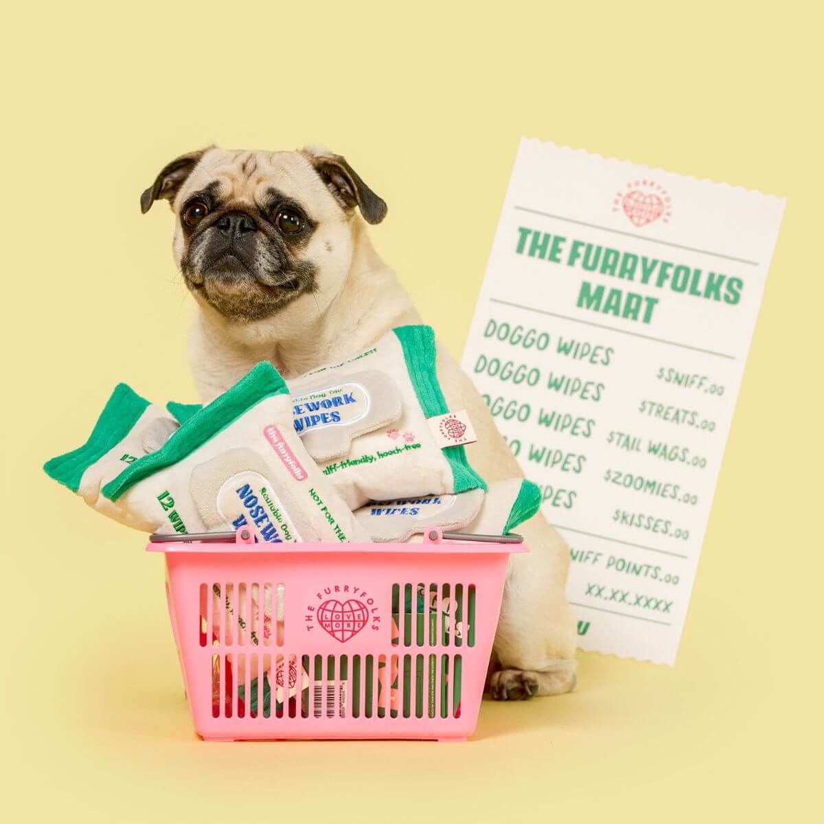 A pug standing behind the pink basket that is filled with the furryfolks doggo wipe nosework toys. 