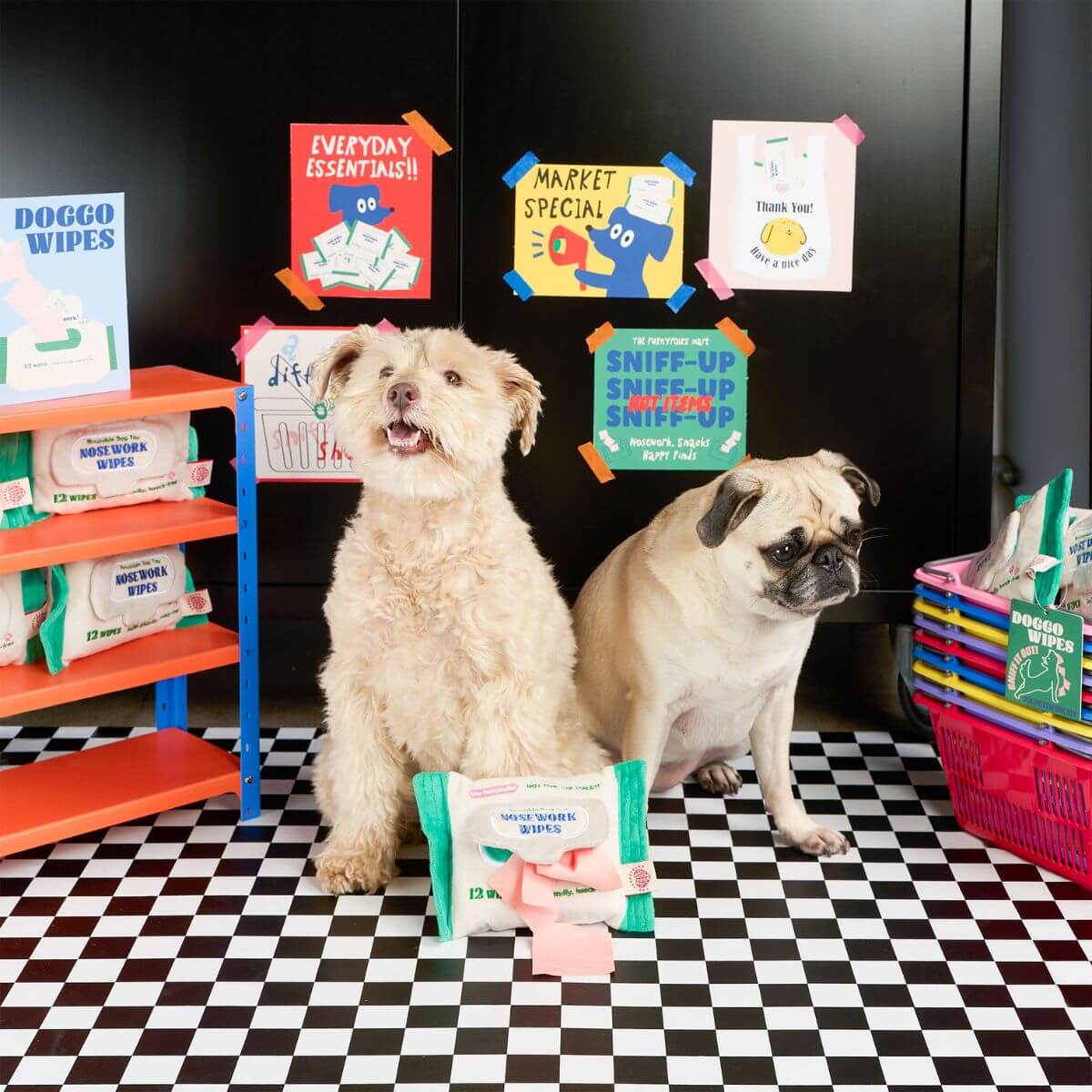 2 dogs sitting on checkered black and white tiles and the the furryfolks doggo wipe nosework toy propped up in front of them. 